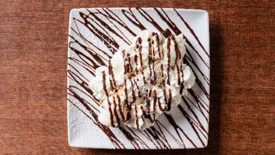 Two cannolis topped with whipped cream, chocolate syrup, and powdered sugar on a white plate.